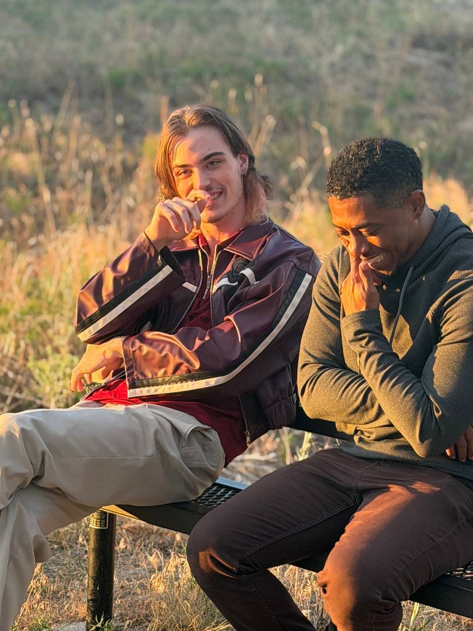 Two actors laughing together on a park bench at sunset